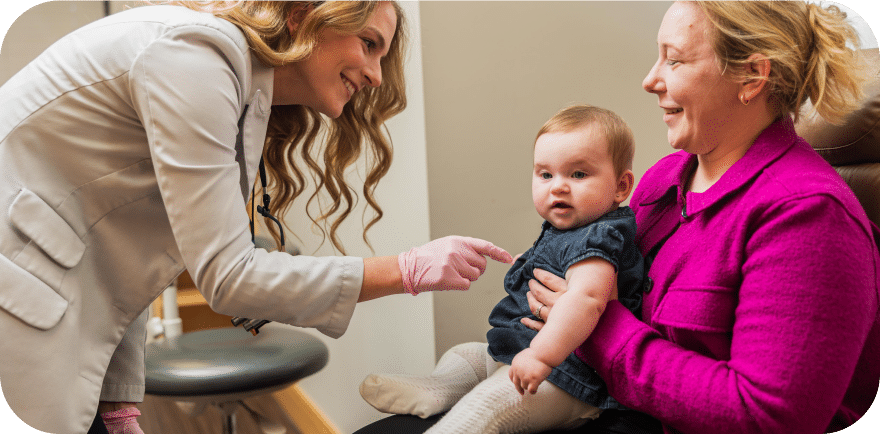 A woman holds a baby as a doctor conducts an examination, showcasing a moment of care and medical attention.