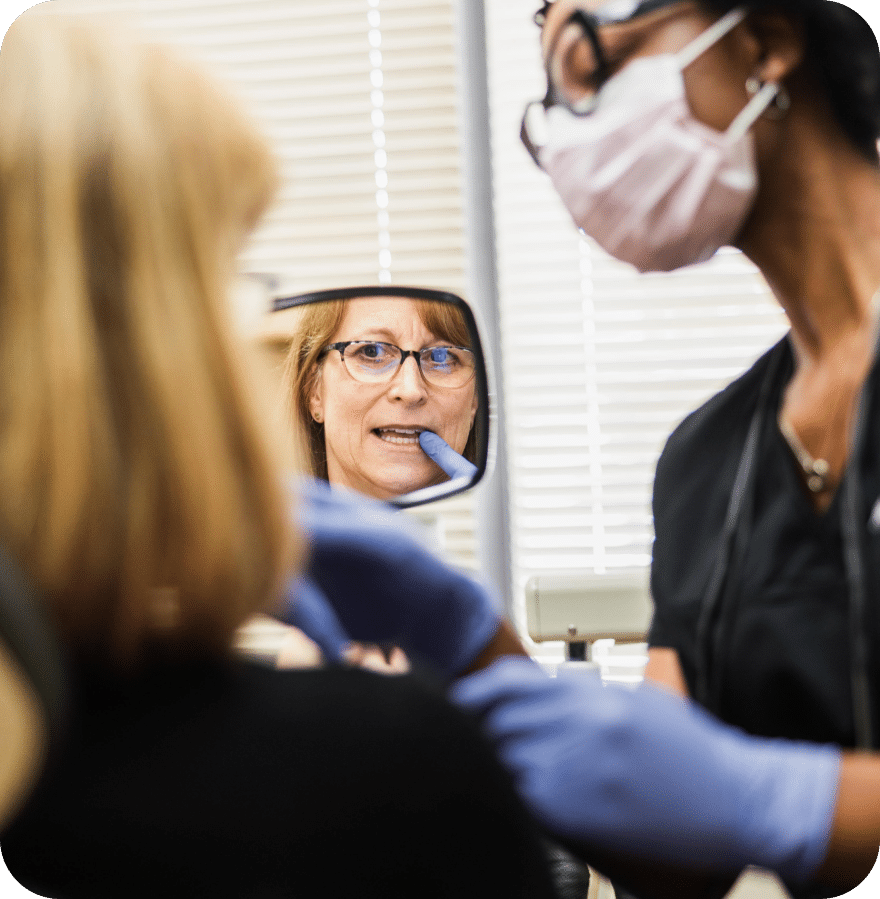 A woman gazes at her reflection in a mirror as a dentist observes her attentively.