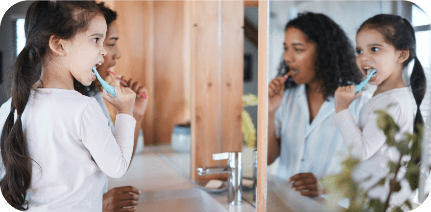 A woman and a little girl are both brushing their teeth, showcasing a moment of shared daily hygiene routine.