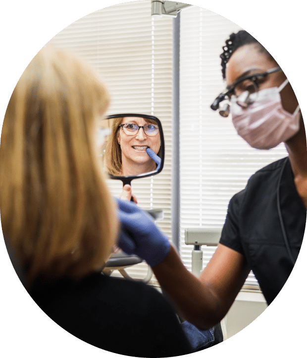 A woman examines her teeth in a dental office using a mirror, showcasing a professional and clean environment.